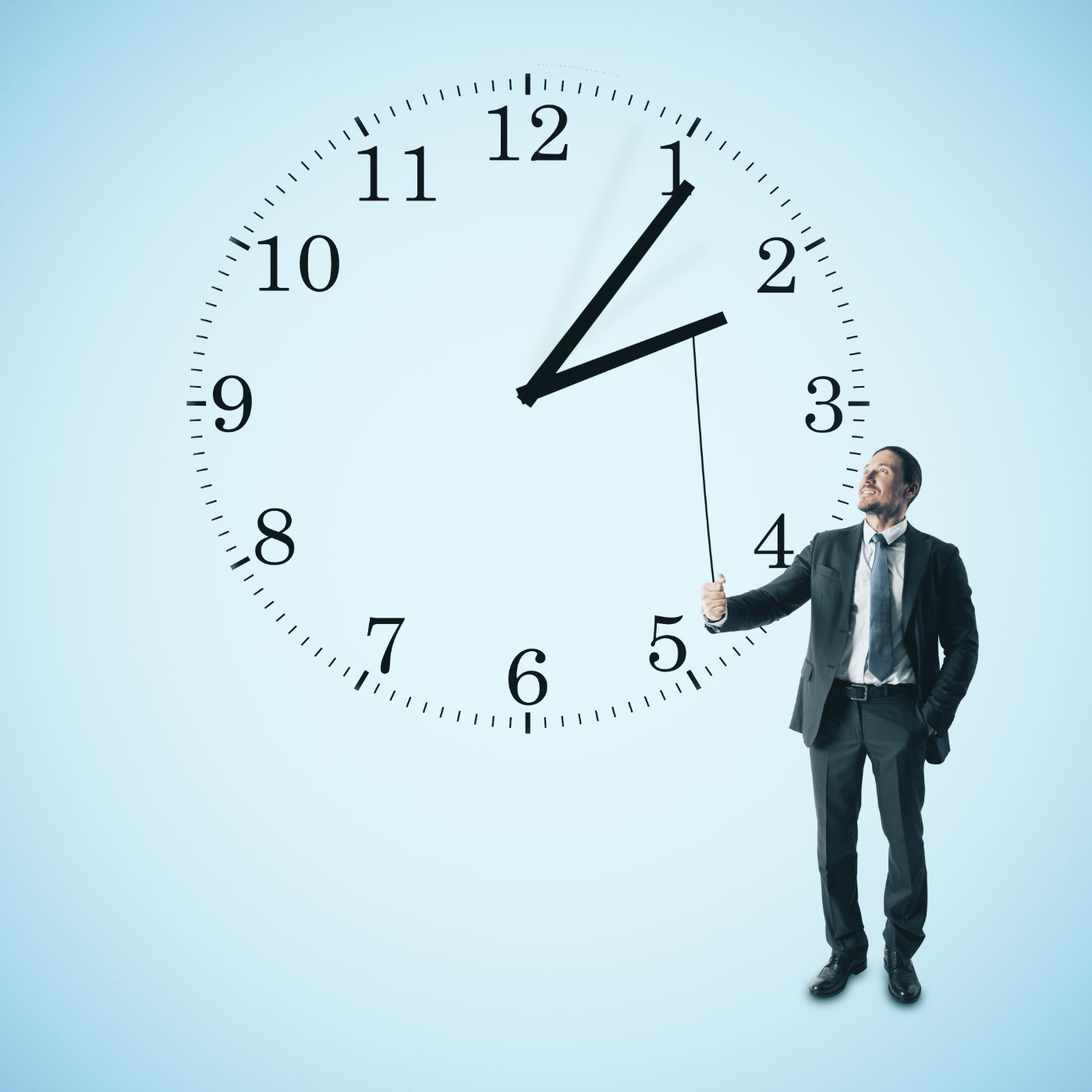 A smiling businessman pulling the hand of a giant clock on a pale sky-blue background