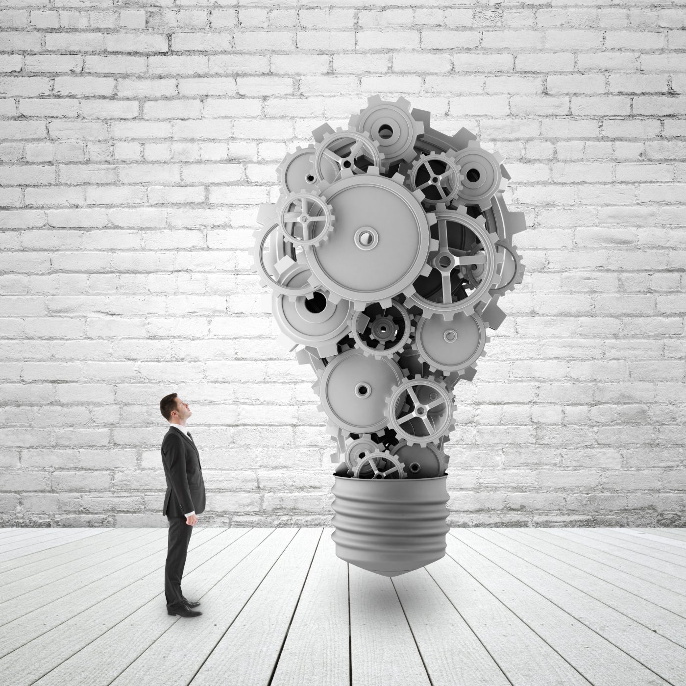 A businessman looking up at a giant light bulb made of grey gears in front of a white brick wall